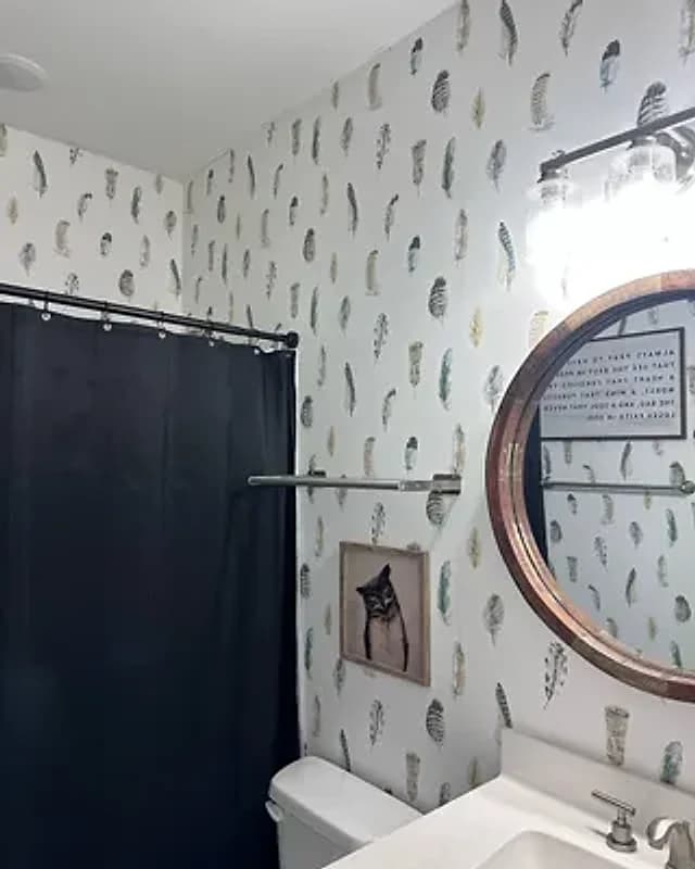Bathroom papered in a white feather-print wallpaper, with a round wood-framed mirror over the sink and a dark shower curtain