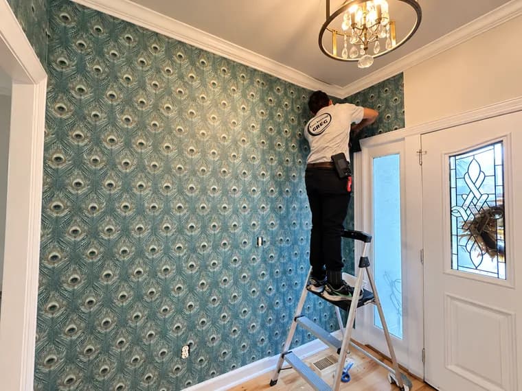 Installer on a step ladder smoothing peacock-pattern wallpaper along the ceiling line of an entryway